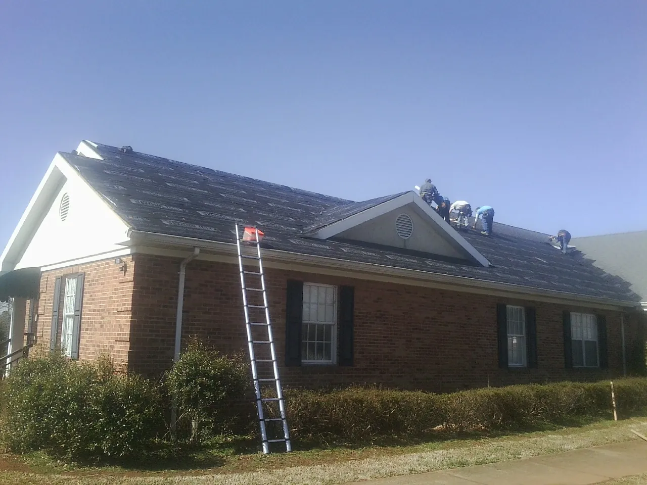 Crew tearing shingles off a brick ranch roof with a ladder set — Emergency Roof Repair work by our crew in Iota
