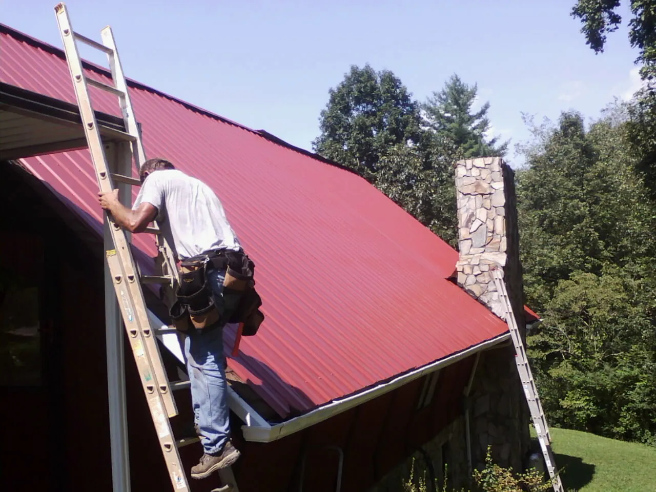Roofer on a ladder beside a red metal roof and stone chimney — Soffit & Fascia Repair by Roofing Brothers in Iota